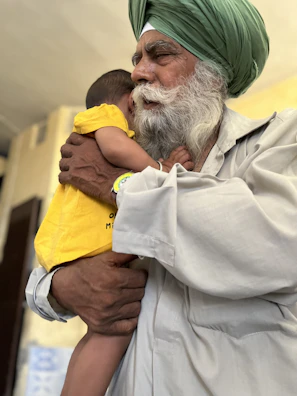 An elderly man with a white beard and a green turban gently holds a young child who is wearing a bright yellow shirt. The man’s expression is calm and tender as he embraces the child, providing a sense of warmth and protection. The background is softly blurred, emphasizing the connection between the two.