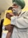 An elderly man with a white beard and a green turban gently holds a young child who is wearing a bright yellow shirt. The man’s expression is calm and tender as he embraces the child, providing a sense of warmth and protection. The background is softly blurred, emphasizing the connection between the two.