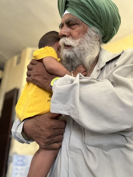 An elderly man with a white beard and a green turban gently holds a young child who is wearing a bright yellow shirt. The man’s expression is calm and tender as he embraces the child, providing a sense of warmth and protection. The background is softly blurred, emphasizing the connection between the two.