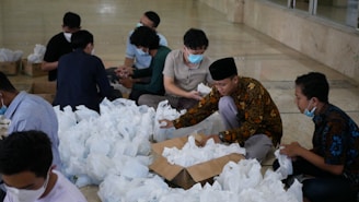 Several people are sitting on the floor, organizing and packing a large number of white bags, which appear to contain items such as food or supplies. The individuals are wearing masks and are focused on their task, with some wearing traditional clothing.
