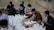Several people are sitting on the floor, organizing and packing a large number of white bags, which appear to contain items such as food or supplies. The individuals are wearing masks and are focused on their task, with some wearing traditional clothing.