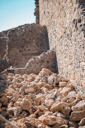A pile of irregularly shaped rocky stones is situated in front of a rough-textured stone wall under clear, bright daylight.
