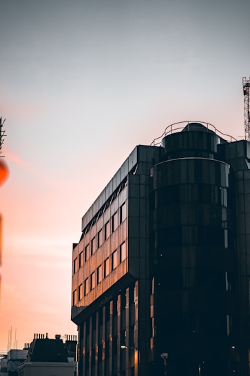 The image captures a modern multi-story building with a glass facade reflecting the light of a sunset. The architecture features sleek lines and a cylindrical section, possibly with balconies or observation points. The sky is a blend of soft colors, transitioning from warm pinks to a muted gray, creating a calming backdrop.