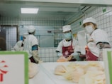 Several individuals wearing white uniforms and face masks are working in a kitchen environment. They are preparing food, with some pastries visible in the foreground and a tiled wall background.