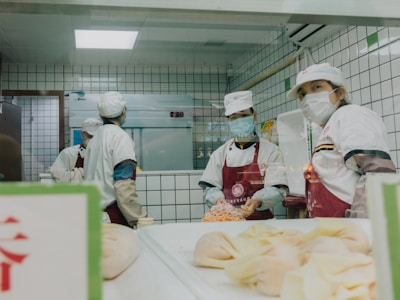 Several individuals wearing white uniforms and face masks are working in a kitchen environment. They are preparing food, with some pastries visible in the foreground and a tiled wall background.