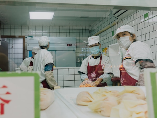 Several individuals wearing white uniforms and face masks are working in a kitchen environment. They are preparing food, with some pastries visible in the foreground and a tiled wall background.
