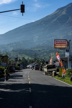 A winding road leads into the distance with a lush mountain backdrop under clear blue skies. On the right, there are stores and vehicles parked, including buses and motorcycles. The road is flanked with various signages and flags, one of which is prominently displaying the word 'Alfamart'. The surrounding landscape features a green, hilly terrain.