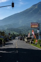 A winding road leads into the distance with a lush mountain backdrop under clear blue skies. On the right, there are stores and vehicles parked, including buses and motorcycles. The road is flanked with various signages and flags, one of which is prominently displaying the word 'Alfamart'. The surrounding landscape features a green, hilly terrain.