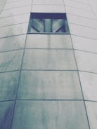 The image features a portion of a modern building facade made up of large, rectangular metal panels with a small, dark rectangular window near the top. The panels are slightly reflective and have a brushed texture, giving a sleek and contemporary appearance.