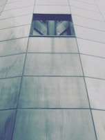The image features a portion of a modern building facade made up of large, rectangular metal panels with a small, dark rectangular window near the top. The panels are slightly reflective and have a brushed texture, giving a sleek and contemporary appearance.