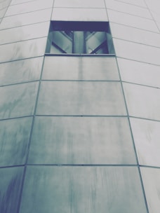 The image features a portion of a modern building facade made up of large, rectangular metal panels with a small, dark rectangular window near the top. The panels are slightly reflective and have a brushed texture, giving a sleek and contemporary appearance.