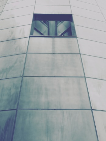 The image features a portion of a modern building facade made up of large, rectangular metal panels with a small, dark rectangular window near the top. The panels are slightly reflective and have a brushed texture, giving a sleek and contemporary appearance.