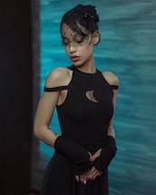 A person wearing an elegant black outfit with unique shoulder cutouts stands against a serene, bluish background. The individual has a delicate black veil with a polka dot pattern partially covering their face and hair. They are posed with hands gently clasped in front, accentuated by dark arm coverings and distinctive pink and white painted nails.