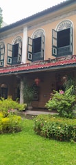 A two-story building with arched windows and open wooden shutters. The facade is painted in pastel colors with intricate detailing around the windows. Below the windows, lanterns hang above a porch surrounded by lush greenery and flowering plants. The scene includes a well-maintained garden with various plants and flowers.