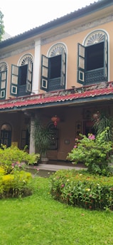 A two-story building with arched windows and open wooden shutters. The facade is painted in pastel colors with intricate detailing around the windows. Below the windows, lanterns hang above a porch surrounded by lush greenery and flowering plants. The scene includes a well-maintained garden with various plants and flowers.