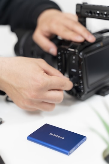 Hands are adjusting the settings on a high-end camera, with a blue Samsung external hard drive placed in the foreground on a white surface.