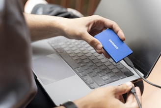 A person holding a blue Samsung external SSD while connecting it to a laptop. The laptop is placed on a table, and the user's focus is on the device connection.