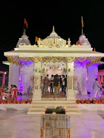 A large, elaborately decorated temple entrance is adorned with colorful lights and garlands. People are gathered around, some seated on red chairs and others standing, suggesting a festive or ceremonial occasion. The architecture features intricate carvings and religious symbols.