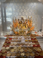 Intricate statues of deities are adorned in vibrant orange and gold garments with floral garlands. In front of them, an array of bowls contains various food offerings, including grains, nuts, and traditional snacks, all arranged on a red cloth. The backdrop features decorative gold patterns on a white surface, creating a reverent and festive atmosphere.