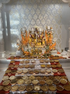 Intricate statues of deities are adorned in vibrant orange and gold garments with floral garlands. In front of them, an array of bowls contains various food offerings, including grains, nuts, and traditional snacks, all arranged on a red cloth. The backdrop features decorative gold patterns on a white surface, creating a reverent and festive atmosphere.