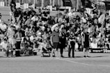 A large crowd of people is gathered, with some standing and others sitting on bleachers. Several individuals are wearing sports attire, and there is a field visible in the foreground with a marker indicating 50 meters. The scene appears to be a sporting event, with a diverse group of spectators.