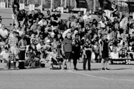 A large crowd of people is gathered, with some standing and others sitting on bleachers. Several individuals are wearing sports attire, and there is a field visible in the foreground with a marker indicating 50 meters. The scene appears to be a sporting event, with a diverse group of spectators.