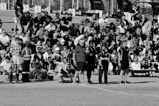 A large crowd of people is gathered, with some standing and others sitting on bleachers. Several individuals are wearing sports attire, and there is a field visible in the foreground with a marker indicating 50 meters. The scene appears to be a sporting event, with a diverse group of spectators.