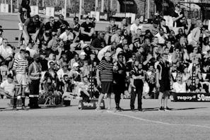 A large crowd of people is gathered, with some standing and others sitting on bleachers. Several individuals are wearing sports attire, and there is a field visible in the foreground with a marker indicating 50 meters. The scene appears to be a sporting event, with a diverse group of spectators.