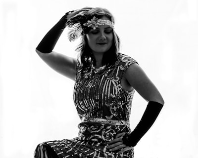 A woman dressed in vintage 1920s fashion, featuring a sequined dress, feathered headband, and long gloves, poses elegantly with one hand on her hip and the other holding the headband.