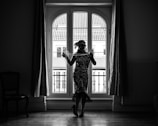 A silhouetted woman dressed in a vintage, flapper-style dress stands indoors in front of a large window with arched panes. Her attire includes a headband and layers of necklaces, and she holds a string of pearls in one hand. The window behind her reveals a view of buildings with tiled rooftops. The room is fairly dark, highlighting the detailed patterns of her clothing against the bright window.