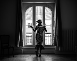 A silhouetted woman dressed in a vintage, flapper-style dress stands indoors in front of a large window with arched panes. Her attire includes a headband and layers of necklaces, and she holds a string of pearls in one hand. The window behind her reveals a view of buildings with tiled rooftops. The room is fairly dark, highlighting the detailed patterns of her clothing against the bright window.