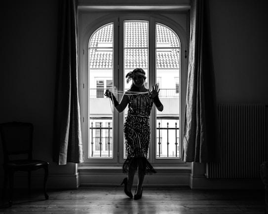 A silhouetted woman dressed in a vintage, flapper-style dress stands indoors in front of a large window with arched panes. Her attire includes a headband and layers of necklaces, and she holds a string of pearls in one hand. The window behind her reveals a view of buildings with tiled rooftops. The room is fairly dark, highlighting the detailed patterns of her clothing against the bright window.