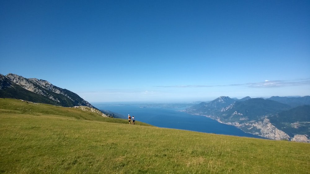 a couple people walking on a grassy hill by a body of water