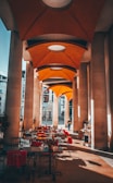 A covered outdoor patio area with tables and chairs is adorned with bright red cushions. Tall, modern columns line the space, supporting an architecturally intriguing orange canopy ceiling. Natural light casts shadows on the floor while a solitary person walks in the background. Some plants are strategically placed around the patio, adding a touch of nature.