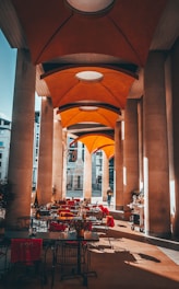 A covered outdoor patio area with tables and chairs is adorned with bright red cushions. Tall, modern columns line the space, supporting an architecturally intriguing orange canopy ceiling. Natural light casts shadows on the floor while a solitary person walks in the background. Some plants are strategically placed around the patio, adding a touch of nature.
