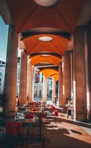 A covered outdoor patio area with tables and chairs is adorned with bright red cushions. Tall, modern columns line the space, supporting an architecturally intriguing orange canopy ceiling. Natural light casts shadows on the floor while a solitary person walks in the background. Some plants are strategically placed around the patio, adding a touch of nature.