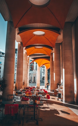 A covered outdoor patio area with tables and chairs is adorned with bright red cushions. Tall, modern columns line the space, supporting an architecturally intriguing orange canopy ceiling. Natural light casts shadows on the floor while a solitary person walks in the background. Some plants are strategically placed around the patio, adding a touch of nature.