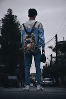 A person wearing a casual blue and white sweater and jeans stands on a street holding a cap in one hand. They are carrying a camouflage backpack and wearing white sneakers. The scene is set in an urban environment with trees and cars visible in the background under a cloudy sky.