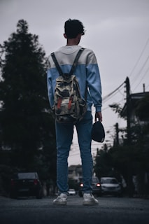 A person wearing a casual blue and white sweater and jeans stands on a street holding a cap in one hand. They are carrying a camouflage backpack and wearing white sneakers. The scene is set in an urban environment with trees and cars visible in the background under a cloudy sky.