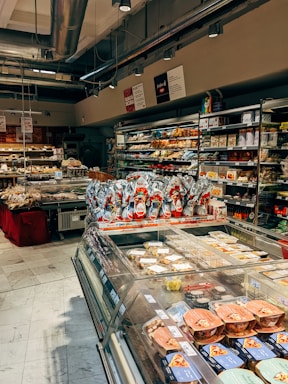 A well-stocked supermarket aisle features shelves filled with various packaged foods, including chocolates and snacks. Refrigerated displays contain assorted prepared meals and desserts. Overhead lights and exposed ductwork give the space an industrial feel.