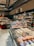 A well-stocked supermarket aisle features shelves filled with various packaged foods, including chocolates and snacks. Refrigerated displays contain assorted prepared meals and desserts. Overhead lights and exposed ductwork give the space an industrial feel.