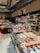 A well-stocked supermarket aisle features shelves filled with various packaged foods, including chocolates and snacks. Refrigerated displays contain assorted prepared meals and desserts. Overhead lights and exposed ductwork give the space an industrial feel.