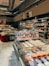 A well-stocked supermarket aisle features shelves filled with various packaged foods, including chocolates and snacks. Refrigerated displays contain assorted prepared meals and desserts. Overhead lights and exposed ductwork give the space an industrial feel.