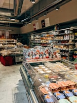 A well-stocked supermarket aisle features shelves filled with various packaged foods, including chocolates and snacks. Refrigerated displays contain assorted prepared meals and desserts. Overhead lights and exposed ductwork give the space an industrial feel.