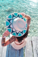 A woman sitting on a wooden pier, wearing a wide-brimmed hat and a Beachcraft tote beside her.