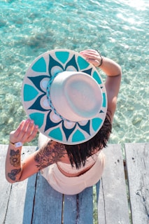 A woman sitting on a wooden pier, wearing a wide-brimmed hat and a Beachcraft tote beside her.