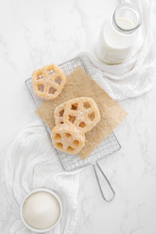 Three sugar-coated pastries, resembling waffle cookies, are placed on a cooling rack lined with brown parchment paper. A bottle of milk is nearby, along with a white cloth and a bowl containing granulated sugar. The background features a white marble surface, creating a clean and minimalistic setting.