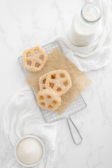 Three sugar-coated pastries, resembling waffle cookies, are placed on a cooling rack lined with brown parchment paper. A bottle of milk is nearby, along with a white cloth and a bowl containing granulated sugar. The background features a white marble surface, creating a clean and minimalistic setting.