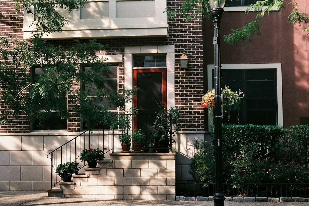A brick rowhouse front with stone steps, a red door and potted plants beside the railing.