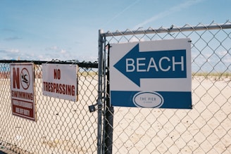 A chain-link fence with several signs is displayed. One sign in red and white reads 'No Swimming When Lifeguards Are Off Duty' with an icon of a swimmer. Another sign says 'No Trespassing' in red letters. A blue sign with a large arrow indicates the direction of the beach, labeled 'The Pier at Seaside.' Behind the fence, there is sand and a glimpse of the ocean under a clear blue sky.